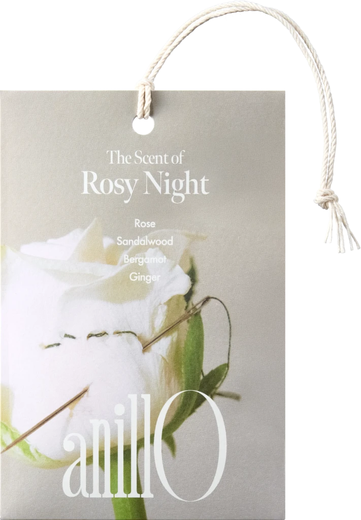 Beige gift tag with a white rope handle reading 'The Scent of Rosy Night' and listing notes: Rose, Sandalwood, Bergamot, Ginger, with a floral image in the background
