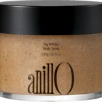 Close-up of a jar with a black lid containing Fig Whisky Body Scrub; beige, speckled scrub visible.