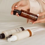 Close-up of hands holding a brown spray bottle with a pump, with other skincare tubes lying on a white surface in the background.