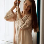Woman in pale pink satin pajamas applying hair product from a brown pump bottle beside a mirror in a bright room.