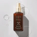 Brown glass bottle with a gold pump and clear cap, labeled Curly Shyll Babassu Oil for Hair on a light background.