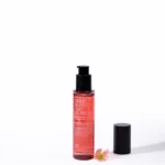 Red cylindrical bottle with black pump dispenser on a white surface, cap off and a small pink flower nearby.