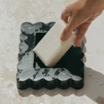Close-up of a hand peeling a white soap slab from a dark, wavy-edged mold with a wet surface nearby.