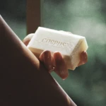 Close-up of a hand holding a white soap bar embossed with 'CORPUS' against a blurred green background.