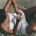 Top-down view of a person in a white shirt applying lotion to their forearm while seated in a woven wicker chair.