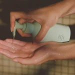 Hands holding a light teal pump bottle and dispensing clear liquid onto the other hand, with wet fingers visible.