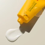 Close-up of a yellow tube dispensing white cream onto a fingertip on a beige background.