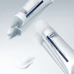 Close-up of white cosmetic tubes with blue labeling on a pale reflective surface, suggestive of skincare cream packaging