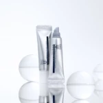 Two metallic silver skincare tubes and a pump bottle arranged on a reflective surface with soft white spheres in the background, cosmetics theme.