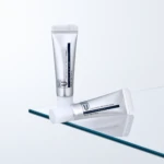 Two silver cosmetic tubes stand upright on a glass surface against a pale blue background, labels partially visible from the side.