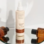 Beige spray bottle labeled Curly Shyll Primer for Damaged Hair on a glass surface with brown dropper bottles nearby.