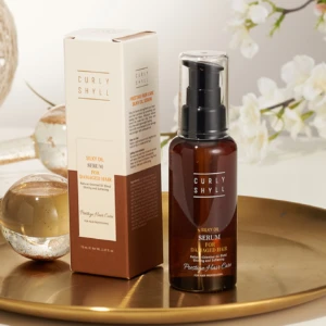 Brown glass serum bottle with a pump next to its cream box that reads 'Curly Shyll Silky Oil Serum for Damaged Hair' on a gold tray with white flowers in the background.
