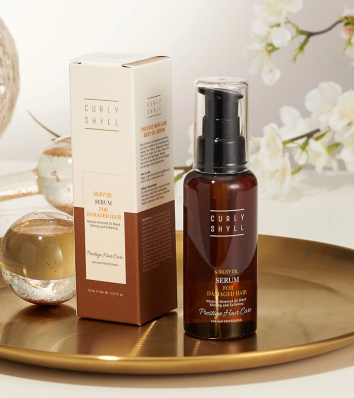 Brown glass serum bottle with a pump next to its cream box that reads 'Curly Shyll Silky Oil Serum for Damaged Hair' on a gold tray with white flowers in the background.