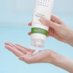 Hand squeezing a white tube labeled 'Curly Shyll' over an open palm, cream being dispensed from the nozzle (hair treatment product)