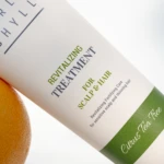 Close-up of a white bottle with green label reading 'Revitalizing Treatment for scalp & hair' placed beside an orange on a light surface.