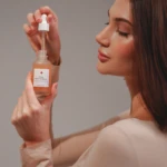 Side profile of a woman with long dark hair applying skincare serum from a dropper bottle held near her face.
