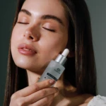 Close-up of a woman's face with eyes closed as she applies a skincare dropper to her neck with a gray hyaluronic serum bottle nearby.