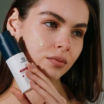 Close-up of a woman applying skincare product to her cheek from a white bottle with a blue cap.