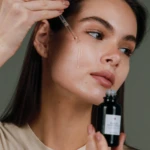 Woman applies facial serum to her cheek using a glass dropper from a dark bottle nearby
