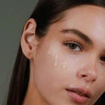 Close-up of a woman's face, showing her left eye, cheek, and lips with streaks of water running down her skin.
