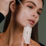 Woman applies clear facial serum to her cheek using a dropper, with a white bottle in view.
