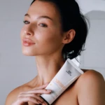 Woman with bare shoulders holds a white facial cleanser tube against her chest, against a neutral background.