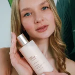 Young woman with light brown hair holding a white bottle of toner close to her face, smiling at the camera.
