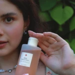 Close-up of a woman applying toner from a pump bottle outdoors, label reads 'Bifida Barrier Toner' with green leaves in the background.