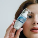 Close-up of a woman holding a glass bottle of vitamin C serum against her cheek, with a dropper top visible.