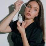 Woman applies skincare serum under her eye using a dropper from a frosted bottle on a white background.