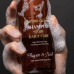 Close-up of a hand holding a brown shampoo bottle with white lather on and around it; label reads Moisture Calm Shampoo for Daily Care.