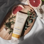 Flat-lay with a beige hair-care tube on a wooden board, next to grapefruit halves, rosemary, and green leaves; product text visible on tube