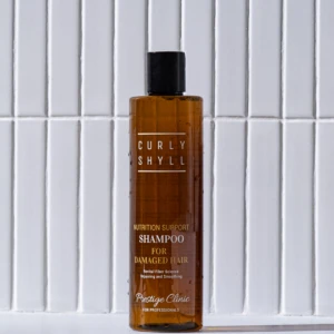 Brown shampoo bottle with black cap standing on a white surface in front of a white tiled wall, label reads 'Nutrition Support Shampoo for Damaged Hair'.