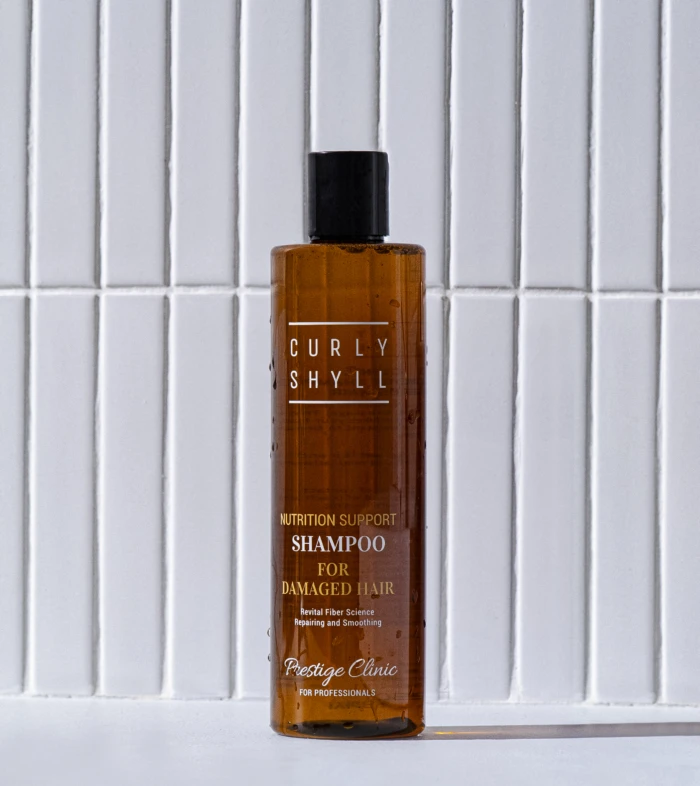 Brown shampoo bottle with black cap standing on a white surface in front of a white tiled wall, label reads 'Nutrition Support Shampoo for Damaged Hair'.