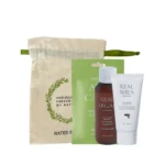 Beige linen drawstring bag with a green ribbon beside a haircare set: a green sachet, a brown argan oil shampoo bottle, and a white Real Shea leave‑in treatment tube.